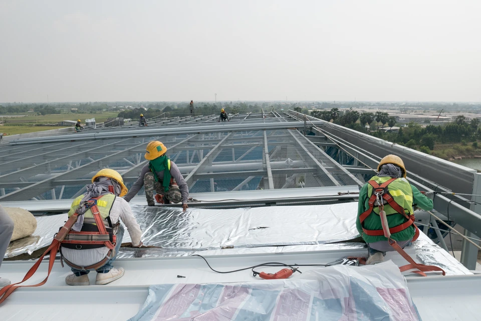 Chantier de construction en extérieur, ouvriers en équipement de protection individuelle installant une membrane d'étanchéité sur toiture-terrasse